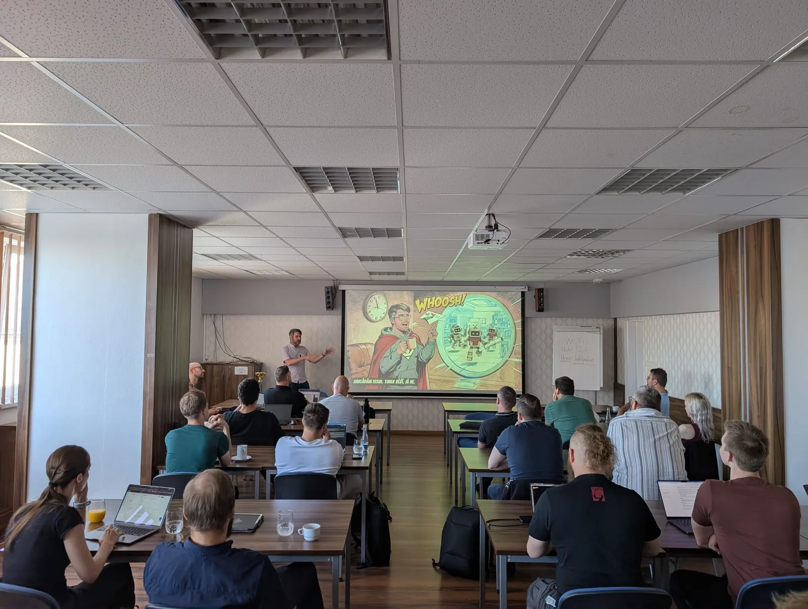 Jiří George Dolejš přednáší na AI BOOTCAMPu v Táboře, na plátně komiksový slide, sál plný účastníků s notebooky